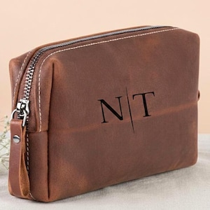 May include: A brown leather toiletry bag with a silver zipper and a leather pull tab. The bag has the initials "NT" printed on the front. The bag is sitting on a white surface.