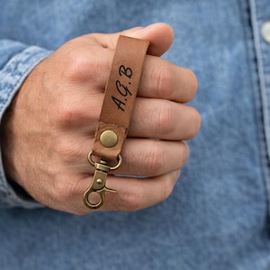 May include: A brown leather keychain with the initials "A.G.B" engraved in black. The keychain has a brass-colored clasp and a snap closure. The leather is a warm, medium brown color.