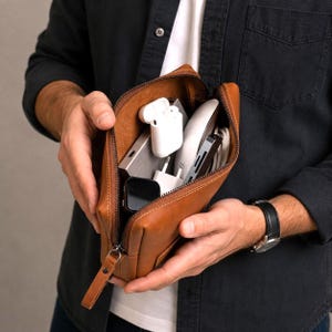 May include: A tan leather zippered pouch filled with various electronic accessories, including white wireless earbuds, a silver power bank, a white wall charger, a black smartwatch, and a multi-port adapter with a white cable.