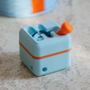 May include: A light blue and orange 3D-printed cube with a lever. The cube has an orange stripe around the middle and a matching orange lever. The design features a slot and a hole on the side.