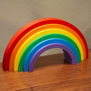 May include: A colorful rainbow-shaped decorative object. The rainbow features bands of red, orange, yellow, green, blue, and purple. The rainbow is made of a solid material and sits on a wooden surface. The rainbow is a decorative item.