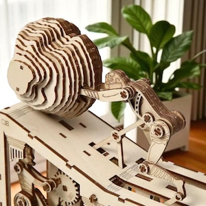 Puede incluir: Una escultura cinética de madera que representa una figura empujando una esfera en capas por un plano inclinado. La escultura está hecha de madera marrón clara y oscura, con intrincados detalles mecánicos. El diseño muestra una mezcla única de arte e ingeniería.