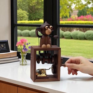 May include: A wooden music box featuring a monkey figurine. The monkey is brown with light-colored accents and sits atop a wooden box with visible gears and a crank. The background includes a window with a view of greenery and flowers.