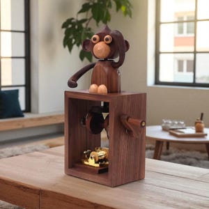 Puede incluir: Una caja de música de madera con una figura de mono. El mono tiene la cara y las orejas de color marrón claro, con el cuerpo y los brazos de color marrón oscuro. La caja es de color marrón oscuro con una manivela y un mecanismo dorado visible en el interior. El mono está posando con la mano en la cabeza.