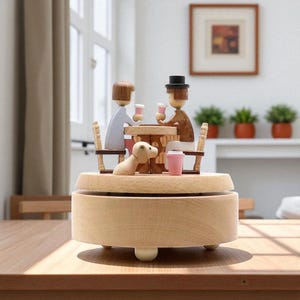 Może przedstawiać: Drewniana pozytywka przedstawiająca miniaturową scenę dwóch postaci przy stole z psem. Postacie trzymają różowe kubki. Pudełko jest jasnobrązowe z okrągłą podstawą i trzema małymi nóżkami.