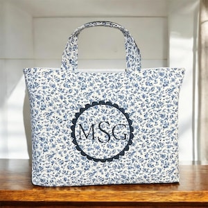May include: A quilted tote bag with a floral pattern in white and blue. The bag features a scalloped circle with the initials "MSG" in the center. The bag has a rectangular shape with a handle.