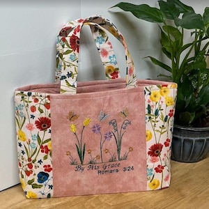 以下が含まれることがあります： 蝶と花が刺繍されたピンク色のセンターパネルが付いたフローラルトートバッグ。バッグは、側面とハンドルにカラフルなフローラルパターンが特徴です。前面のテキストは「By His Grace Romans 3:24」と書かれています。