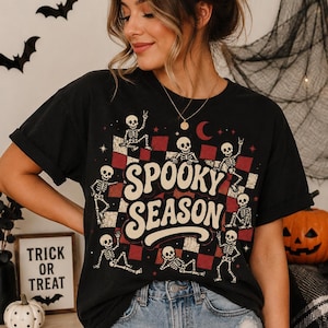 May include: A black t-shirt featuring a graphic print of multiple skeletons in various poses over a red and black checkered background. The text "SPOOKY SEASON" is printed in large, cream-colored, wavy lettering across the center of the design.