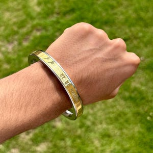 May include: A silver and gold-toned bangle bracelet with engraved text and symbols. The bracelet is worn on a wrist, with a green grassy background. The bangle has a polished finish and a simple, elegant design.