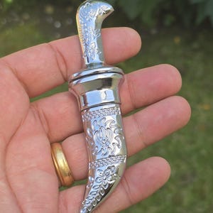 May include: A small, polished silver-coloured metal kirpan with an ornate curved handle and a sheath featuring intricate floral engravings and a central Khanda symbol. The decorative object has a curved blade profile and a reflective metallic finish.
