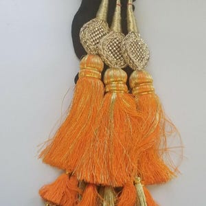 May include: A cluster of decorative tassels featuring vibrant orange and gold accents. The tassels have a black base, with gold-colored, ornate tops and orange fringe. Small gold bells hang from the bottom of the tassels, adding a touch of elegance.
