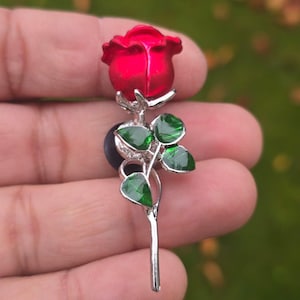 Könnte beinhalten: Rote Rosenbrosche mit grünen Blättern und einem silbernen Stiel. Die Rose ist leuchtend rot und die Blätter sind tiefgrün. Die Brosche ist aus Metall gefertigt und hat eine Nadel auf der Rückseite.