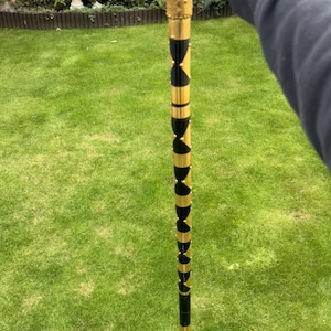 May include: A walking stick featuring a curved handle with a textured surface. The shaft is decorated with a repeating pattern of alternating black and gold segments, accented by small gold studs running vertically along the length of the cane.