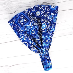May include: A blue paisley patterned headband with white accents. The headband is knotted at the bottom and has an elastic band. The fabric has a repeating pattern of floral and geometric designs.