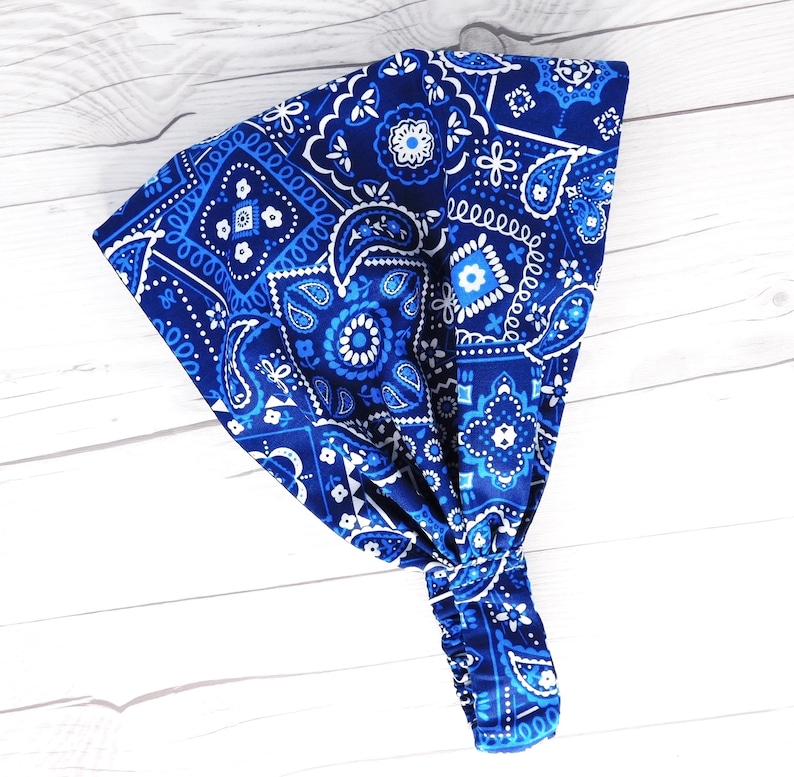 May include: A blue paisley patterned headband with white accents. The headband is knotted at the bottom and has an elastic band. The fabric has a repeating pattern of floral and geometric designs.