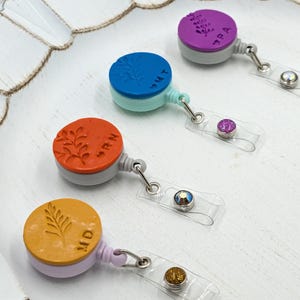 Peut inclure: Quatre porte-badges rétractables colorés avec différents motifs floraux et les mots "TPA", "INT", "ORN" et "MID" dessus. Chaque porte-badge est doté d'une sangle en plastique transparente avec un cristal décoratif.
