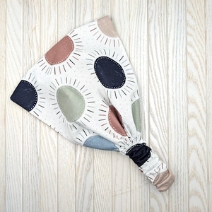 May include: A white fabric headband with a repeating pattern of colorful sunburst designs in shades of blue, brown, and green. The headband has a gathered elastic section and a solid navy blue end.