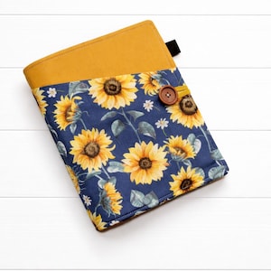 Puede incluir: Una funda de libro de tela con la parte superior de color amarillo mostaza y la inferior azul marino. La parte inferior presenta un estampado de girasoles con flores amarillas y hojas verdes. Un botón de madera asegura la funda.