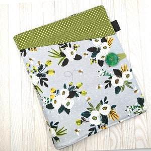 May include: A grey and white fabric book cover with a floral pattern featuring white flowers, green leaves, and yellow bees. The cover has a green polka dot flap and a green button.