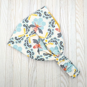 May include: A wide headband with a colorful bee and honeycomb pattern. The design features bees with red and black wings, blue and yellow honeycomb shapes, and paisley elements on a cream background. The headband is made of soft fabric.