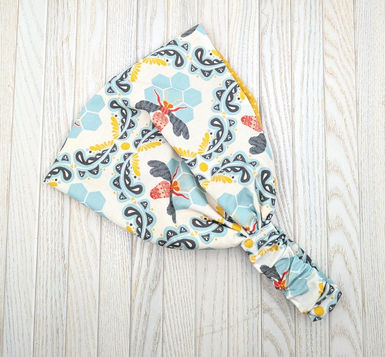May include: A wide headband with a colorful bee and honeycomb pattern. The design features bees with red and black wings, blue and yellow honeycomb shapes, and paisley elements on a cream background. The headband is made of soft fabric.