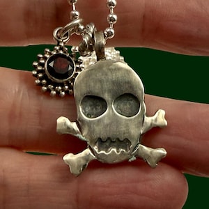 May include: A silver-toned necklace featuring a skull pendant with crossbones and a small sunburst charm with a dark red stone. The necklace has a ball chain design.