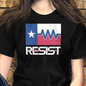 Peut inclure: T-shirt noir avec un motif graphique représentant le drapeau du Texas, une ligne de battement de cœur et le mot "RESIST" en blanc. Le motif utilise les couleurs bleu, blanc et rouge.
