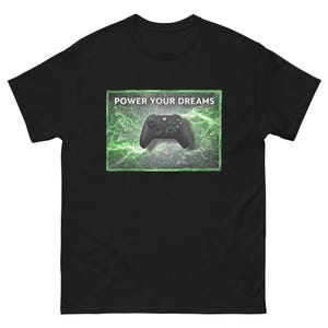 Puede incluir: Camiseta negra con un gráfico de un mando de videojuegos y el texto "POWER YOUR DREAMS". El mando está sobre un fondo verde y gris.