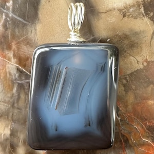 May include: A rectangular pendant featuring a smooth, polished dark gray and translucent blue agate stone. The stone is encased in a silver-toned wire wrap at the top, forming a circular bail for hanging. The surface displays natural banding patterns.