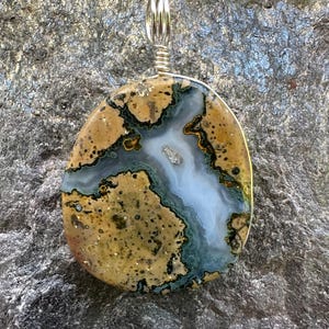 May include: A polished, oval-shaped pendant featuring a natural stone with a tan and brown speckled surface, accented by a central vein of translucent white and deep blue-green mineral deposits, secured by a silver-toned wire bail at the top.
