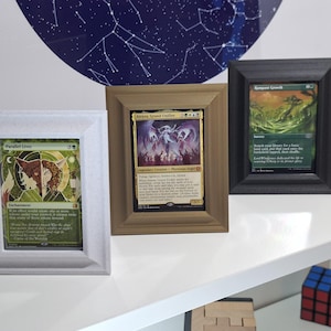 Könnte beinhalten: Drei gerahmte Magic: The Gathering-Karten. Die Rahmen sind weiß, beige und schwarz. Die Karten zeigen Fantasy-Kunst und Text. Die Rahmen werden auf einem weißen Regal ausgestellt. Ein Zauberwürfel befindet sich in der unteren rechten Ecke.