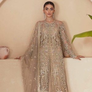 May include: A beige formal gown with intricate embroidery and embellishments. The long-sleeved dress features a sheer neckline and a flowing skirt. A matching sheer shawl is draped over the shoulders. The gown is adorned with floral and geometric patterns.