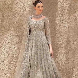 Puede incluir: Un vestido largo y fluido de color gris plateado con intrincados bordados y abalorios. El vestido presenta mangas largas, un cuello alto y un chal a juego. El diseño es adecuado para eventos formales.