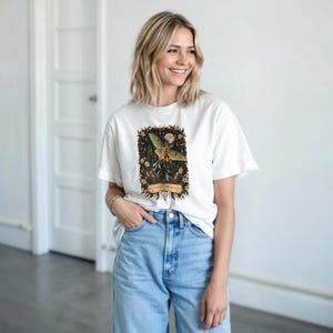 Może przedstawiać: Biały t-shirt z krótkim rękawem z centralną grafiką przedstawiającą zieloną ćmę księżycową otoczoną ciemnymi elementami kwiatowymi i motywami niebieskimi. Pod ćmą znajduje się baner z napisem, "PRETTY THINGS ARE AWESOM".