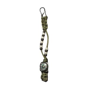Puede incluir: Un llavero de paracord verde con una pequeña brújula y un mosquetón de metal. El llavero presenta una serie de cuentas negras y transparentes. La brújula tiene una carcasa negra y una esfera verde y blanca.