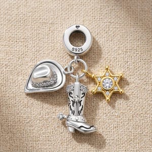 Pode incluir: Um berloque de prata com um chapéu de cowboy, uma bota western e uma estrela de xerife dourada, decorados com pequenas pedras claras. O aro superior tem a gravação S925. Os pingentes estão presos a um anel central de prata.
