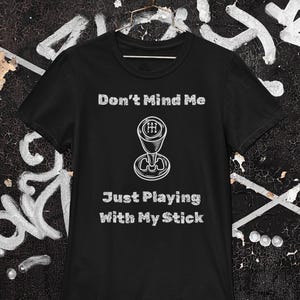 May include: Black t-shirt with the text "Don't Mind Me" above a graphic of a gear shift and the text "Just Playing With My Stick". The shirt is hanging on a metal hanger.