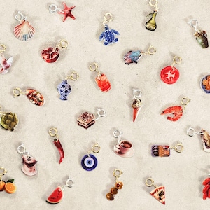 May include: A selection of colourful charms with gold-tone clasps. The charms feature diverse objects, including a seashell, starfish, turtle, bottle, and food items such as a tomato, ice cream cone, and pizza slice. The charms are arranged on a light-coloured surface.