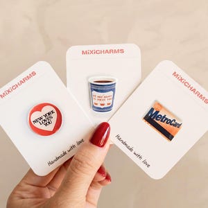 Puede incluir: Tres pines de esmalte sobre tarjetas de soporte blancas etiquetadas como MixiCHARMS. Un pin es un corazón rojo con el texto NEW YORK LOVES YOU, otro es un vaso de café azul y blanco con el texto WE ARE HAPPY TO SERVE YOU, y otro es una MetroCard amarilla y azul.