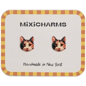 Op de afbeelding: Stud oorbellen met kattengezicht op een crème kaart met een gele en bruine rand. De oorbellen zijn zwart, bruin en wit. De kaart leest "MixiCharms" en "Handmade in New York".