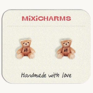 May include: A pair of teddy bear stud earrings on a cream-colored card. The earrings are light brown with black eyes and the text "I ❤️ NY". The card has the text "MixiCharms" and "Handmade with love".