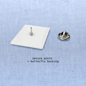 Peut inclure: &Eacute;pinglette rectangulaire blanche avec une tige argent&eacute;e et un fermoir papillon s&eacute;par&eacute;. Le texte "secure posts + butterfly backing" est visible. L'&eacute;pinglette est pr&eacute;sent&eacute;e sur un tissu bleu clair.