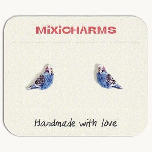 May include: A pair of stud earrings featuring blue and white budgerigar bird designs on a rectangular display card. The card displays the text MIXICHARMS at the top and the phrase Handmade with love at the bottom in black lettering.