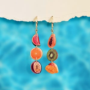 May include: Gold-tone earrings featuring a series of colorful fruit slices. Each earring showcases a watermelon wedge, orange slice, passion fruit half, and a mango. The other earring displays a papaya, kiwi, and mango. The earrings hang against a blue background.