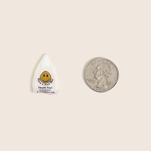 Peut inclure: Un emballage de bonbon blanc, de forme triangulaire, avec un smiley jaune et le texte "Have a Nice Day! Thank You!" &agrave; c&ocirc;t&eacute; d'une pi&egrave;ce de monnaie. L'emballage mesure environ 2,5 cm de long.