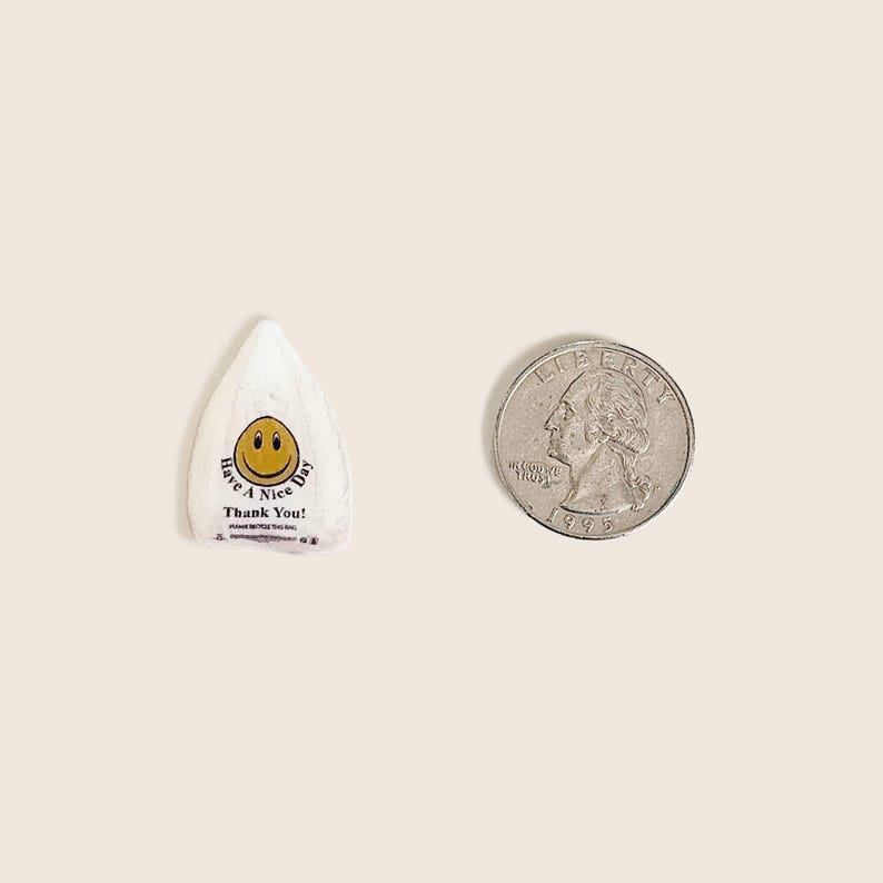 Peut inclure: Un emballage de bonbon blanc, de forme triangulaire, avec un smiley jaune et le texte "Have a Nice Day! Thank You!" &agrave; c&ocirc;t&eacute; d'une pi&egrave;ce de monnaie. L'emballage mesure environ 2,5 cm de long.