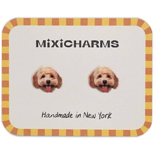 May include: A pair of MixiCharms stud earrings featuring a smiling dog face with a pink tongue. The earrings are on a white card with the text "MixiCharms" and "Handmade in New York". The card has a yellow and orange border.