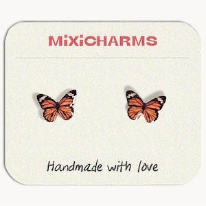 Puede incluir: Un par de pendientes de botón con forma de mariposas monarca naranjas y negras, presentados sobre una tarjeta rectangular de color crema. La tarjeta muestra el texto MIXICHARMS arriba y la frase Handmade with love impresa abajo.