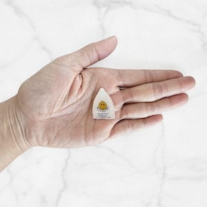 Peut inclure: Une petite &eacute;tiquette blanche en forme de goutte d'eau avec un smiley jaune et le texte "Have a Nice Day! Thank You!" est tenue dans la paume ouverte d'une personne. L'&eacute;tiquette a un sommet arrondi et un fond pointu. Fond blanc marbr&eacute;.