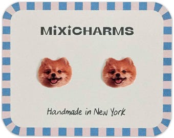 May include: A pair of stud earrings featuring smiling Pomeranian dog faces. The earrings are displayed on a white card with a blue and pink checkered border. The card reads "MixiCharms" and "Handmade in New York".